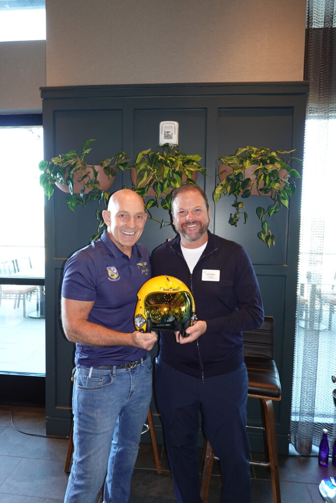 John “Gucci” Foley and Jason Pritzker smile while holding a yellow Blue Angels helmet.